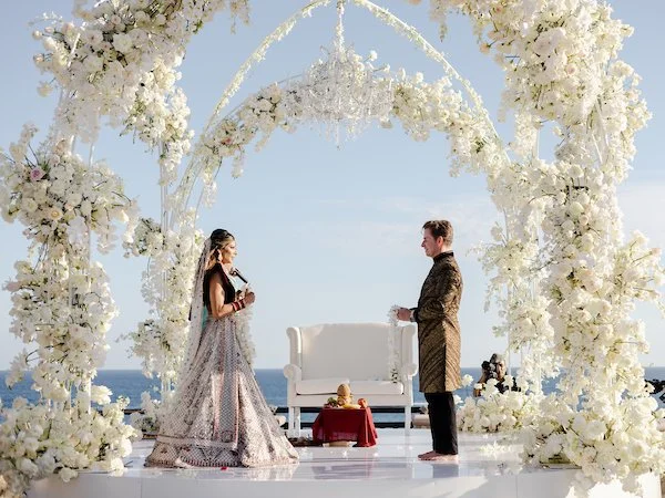 A couple participating in a wedding ceremony outdoors on a decorated stage with white flowers and an arch, against a backdrop of the ocean and sky.
